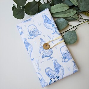 May include: A rectangular, white fabric item with a blue rooster and egg basket pattern. A wooden button and elastic closure are visible. The item is decorated with a farm animal theme.