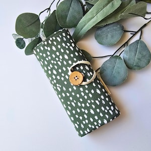 May include: A rolled-up, green fabric case with a white dotted pattern, secured with a beige strap and a wooden button. The case is surrounded by green eucalyptus leaves on a white background.