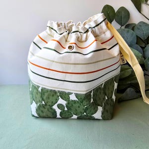 May include: A drawstring bag with a white background, featuring horizontal stripes in orange, green, and brown. The bottom section displays a green cactus print. A yellow strap is attached to the bag.