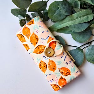 May include: A white fabric pouch with a lemon print and a brown button closure. The pouch has a white strap with the text "Knit & Stitch" printed on it.
