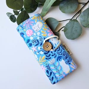 May include: A blue fabric glasses case with a floral and cat pattern. The case features a button closure and a small tag that reads "Knit & Purr". The design includes white cats, pink flowers, and yellow accents.