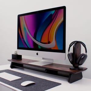 May include: A wooden monitor stand with a black metal frame, holding a silver computer monitor with a colorful abstract background. A pair of black headphones are on a wooden stand to the right of the monitor.