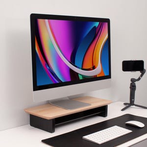 Op de afbeelding: Een zwarte computermonitor met een kleurrijk abstract ontwerp op het scherm staat op een houten standaard met een zwarte basis. De monitor staat op een wit bureau met een zwarte muismat, een wit toetsenbord en een zwarte telefoon op een statief.