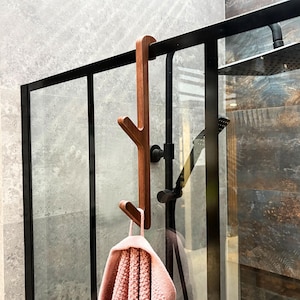 Puede incluir: Un toallero de madera con diseño de rama de árbol cuelga de un marco negro de puerta de ducha. Una toalla rosa cuelga del toallero. La ducha tiene un cabezal de ducha negro y una puerta de cristal. La pared es de baldosas grises y marrones.