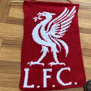 May include: A red and white knitted banner featuring the Liverpool Football Club emblem, a Liver bird. The banner has the letters "L.F.C." at the bottom. A wooden dowel is at the top for hanging.