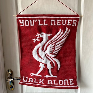May include: Red and white banner with the text "YOU'LL NEVER WALK ALONE" and a white bird emblem. The banner is hanging on a door. The banner is made of a knitted material.