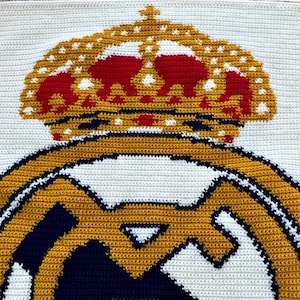 May include: Handmade crochet blanket featuring the Real Madrid logo. The design includes a gold crown with red accents atop a gold and navy emblem, all set against a white background. The blanket is crafted with a tight stitch.
