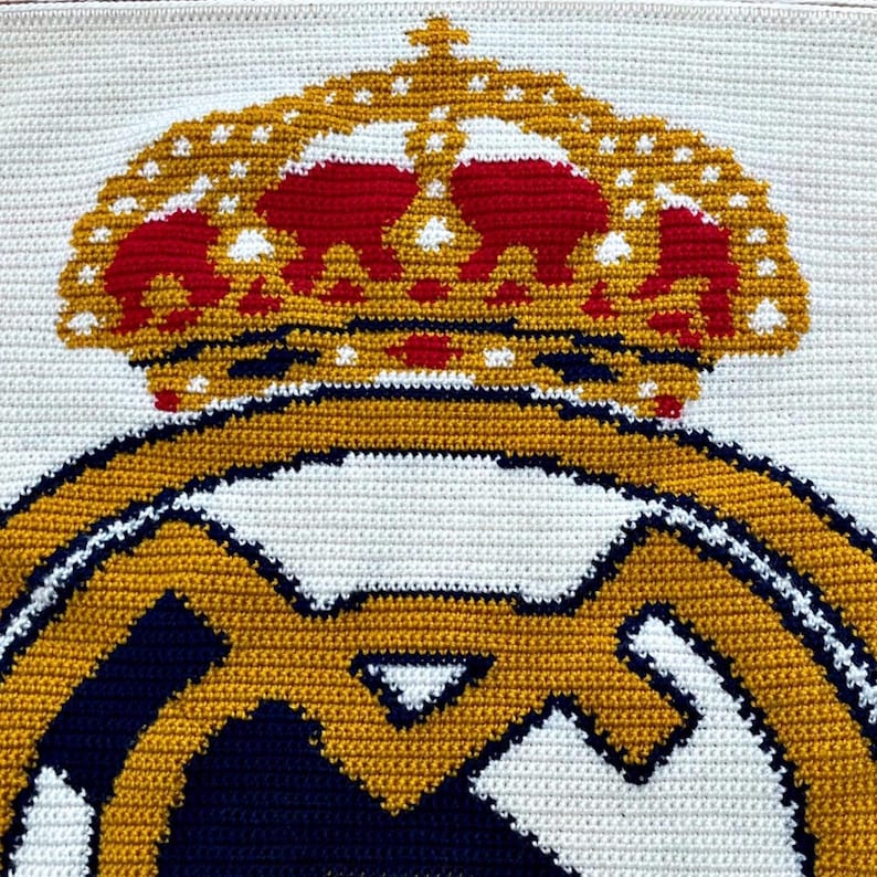 May include: Handmade crochet blanket featuring the Real Madrid logo. The design includes a gold crown with red accents atop a gold and navy emblem, all set against a white background. The blanket is crafted with a tight stitch.