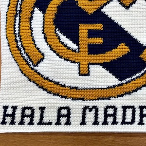 May include: Crocheted blanket featuring the Real Madrid logo in white, navy blue, and gold. The words "HALA MADRID" are stitched in navy blue at the bottom. The blanket is made with a tight stitch pattern.