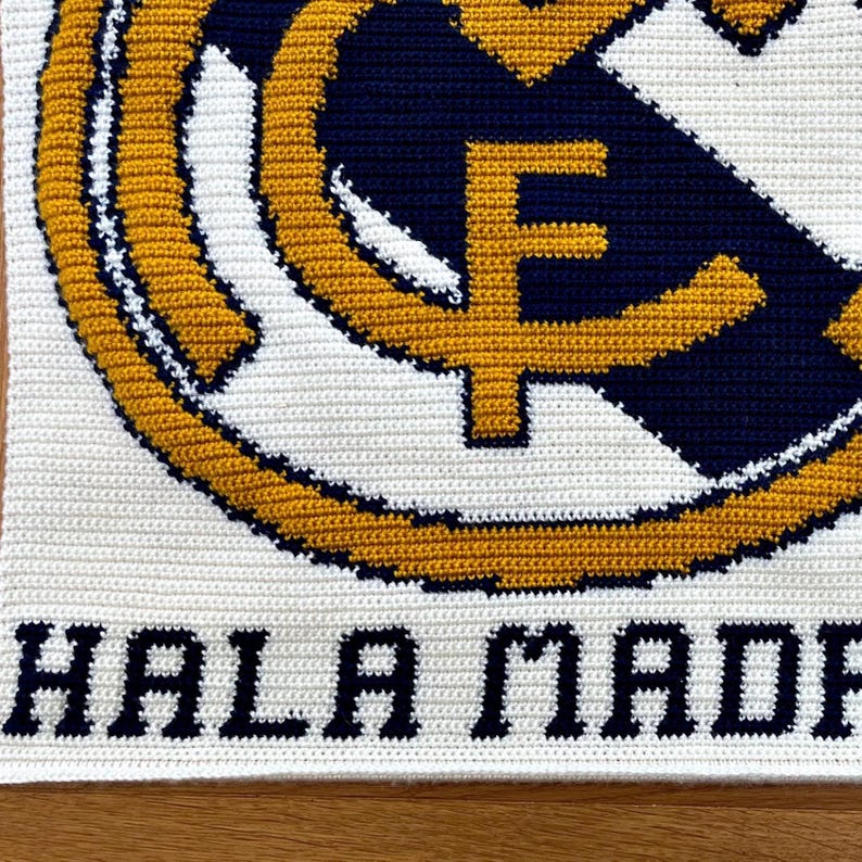 May include: Crocheted blanket featuring the Real Madrid logo in white, navy blue, and gold. The words "HALA MADRID" are stitched in navy blue at the bottom. The blanket is made with a tight stitch pattern.