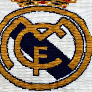 May include: Crocheted emblem featuring the Real Madrid logo. The design includes a gold crown, a gold and navy blue emblem with the letters "R" and "F", and a white background. The emblem is made with a crochet stitch.
