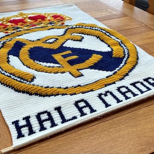 May include: White rectangular textile with the Real Madrid logo in the center. The logo features a gold crown, a gold and navy blue emblem, and the words "HALA MADRID" in navy blue letters.