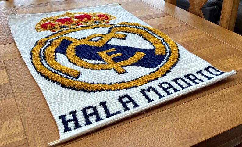 May include: White rectangular textile with the Real Madrid logo in the center. The logo features a gold crown, a gold and navy blue emblem, and the words "HALA MADRID" in navy blue letters.