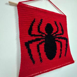 Könnte beinhalten: Ein rechteckiger roter Wandbehang aus Häkelarbeit mit einer großen schwarzen Spinnengrafik in der Mitte. Das Textil ist an einem hellen Holzstab befestigt und verfügt über eine passende rote Garnkordel zum Aufhängen, was ein flaches Display ergibt.