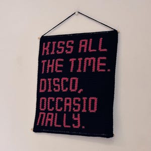 May include: A rectangular black crochet wall hanging featuring the pink text KISS ALL THE TIME. DISCO, OCCASIONALLY. The piece is suspended from a wooden dowel by a black string, creating a textured, handmade display of the quoted phrase.