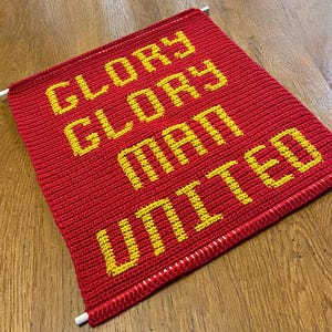 May include: A red knitted banner displaying the text "GLORY GLORY MAN UNITED" in yellow. The banner is rectangular with white dowels at the top and bottom. The text is centrally positioned on the banner.