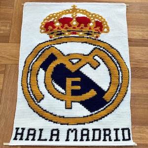 Może przedstawiać: Ręcznie wykonany biały gobelin na ścianę z logo Realu Madryt. Logo jest w kolorze złotym, granatowym i białym, z czerwoną i złotą koroną. U dołu widnieje napis "HALA MADRID".