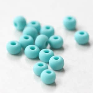 Pu&ograve; includere: Un gruppo di 14 perline rotonde color turchese, sparse su una superficie bianca.