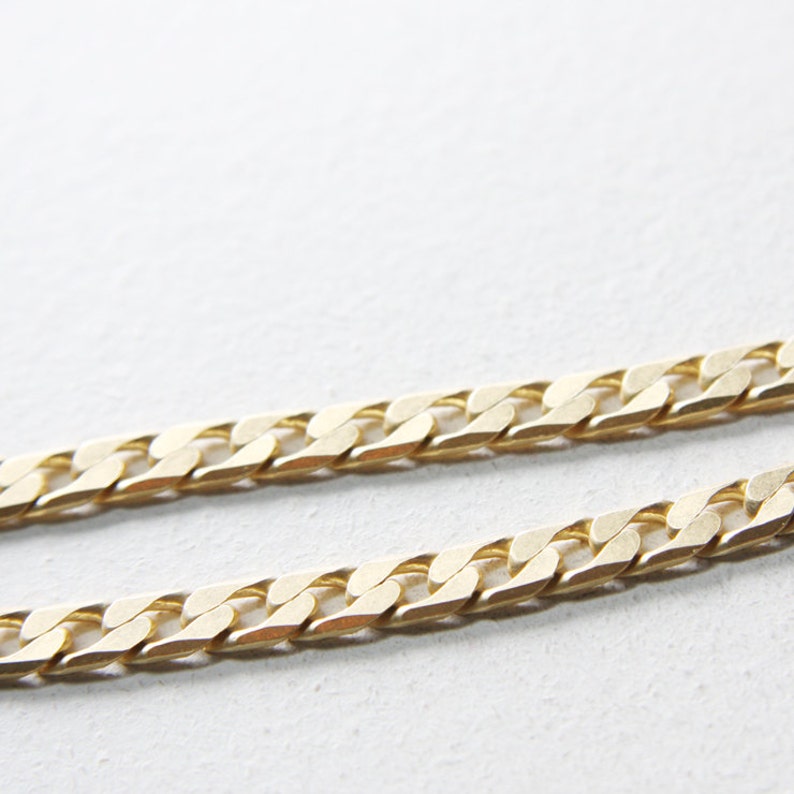 May include: A gold chain necklace with a curb link style. The chain is laying flat on a white surface.