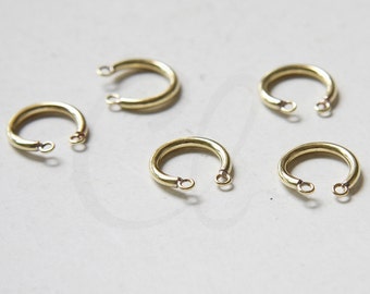 Pode incluir: Cinco anéis de metal de cor dourada com um pequeno laço de cada lado. Os anéis estão dispostos em semicírculo sobre uma superfície branca. Latão bruto.