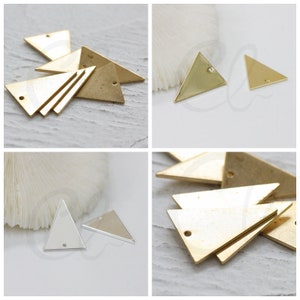 May include: A collection of gold-colored triangle charms with a small hole at the top for jewelry making. The charms are shown in multiple arrangements, including stacked and individual pieces. The metal has a matte finish.