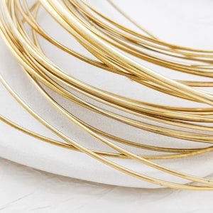 Puede incluir: Primer plano de un haz de alambre brillante de color dorado, enrollado y en capas. El alambre parece liso y de grosor uniforme, descansando sobre una superficie blanca texturizada. La imagen resalta el brillo metálico y la disposición orgánica del alambre.