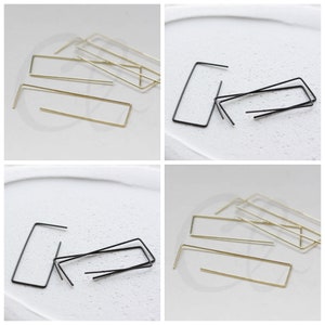 May include: Minimalist earrings in gold and black, featuring a rectangular design with a straight bar and a bent post. The earrings are presented on a white surface. The design is simple and elegant, suitable for everyday wear.