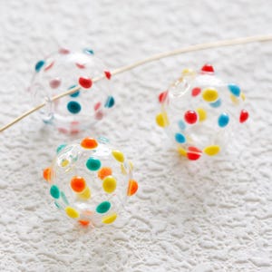 Puede incluir: Tres cuentas de vidrio transparentes con lunares de colores. Las cuentas están enhebradas en un alambre dorado. Las cuentas son rojas, amarillas, azules y verdes.