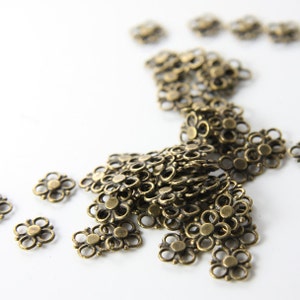 May include: A large pile of antique gold metal flower-shaped charms. The charms are arranged in a loose pile on a white surface.