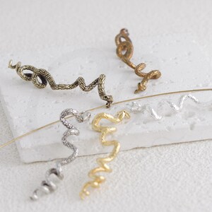Puede incluir: Una colección de piezas de joyería con forma de serpiente en varios acabados metálicos. Las piezas incluyen un brazalete de oreja de tono bronce, un brazalete de oreja de color dorado, un brazalete de oreja de color plateado y un pendiente con forma de serpiente dorada. La joyería se exhibe sobre una superficie blanca.