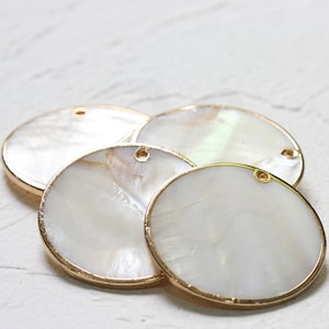 May include: Four round white mother of pearl shell charms with gold trim and a hole for hanging. The charms are stacked on top of each other.
