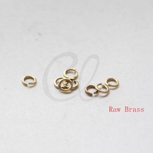 Könnte beinhalten: Nahaufnahme einer Gruppe kleiner, goldfarbener Sprungringe. Die Ringe liegen lose auf einer weißen Oberfläche. Der Text "Raw Brass" ist in Rot unter den Ringen geschrieben.