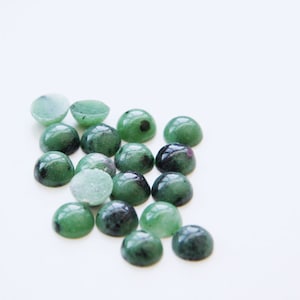 Könnte beinhalten: Eine Sammlung grüner und schwarzer Edelstein-Cabochons, einige mit einem Hauch von Rosa, auf einer weißen Oberfläche angeordnet. Die Cabochons sind rund und haben eine glatte, polierte Oberfläche.