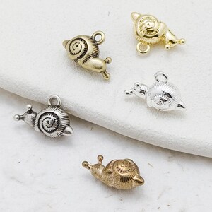 Könnte beinhalten: Eine Sammlung kleiner Metall-Schneckenanhänger in verschiedenen Ausführungen: Gold, Silber und Bronze. Jeder Anhänger hat ein detailliertes Spiralgehäuse und eine Schlaufe zum Befestigen an Schmuck oder anderen Bastelarbeiten. Die Schnecken sind auf einer weißen Oberfläche abgebildet.