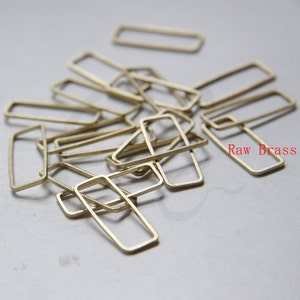 May include: A pile of raw brass rectangular jump rings. The rings are all the same size and shape, and they are scattered randomly on a white surface. The text "Raw Brass" is visible in the lower right corner of the image.