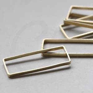 Pode incluir: Close-up de várias molduras retangulares de metal dourado. As molduras são retângulos abertos, com um acabamento fosco. As molduras estão espalhadas sobre uma superfície cinza claro. Estas molduras são ideais para fazer joias.