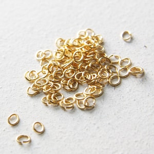 May include: A collection of small, gold-coloured jump rings scattered on a light-coloured surface. These open rings are used for jewellery making and other crafts. The rings are approximately 0.6 cm in diameter.