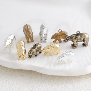 Könnte beinhalten: Eine Sammlung kleiner Bärenanhänger in verschiedenen Metallausführungen, darunter Gold, Silber und Bronze. Die Anhänger sind in verschiedenen Posen, einige stehen und andere gehen. Diese dekorativen Anhänger sind ideal für Schmuckherstellung oder Bastelprojekte.