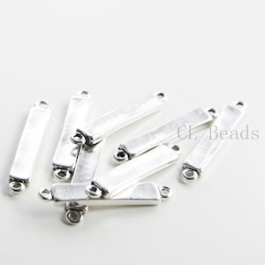 Puede incluir: Un grupo de conectores de barra de metal plateados con dos bucles en cada extremo. Los conectores están dispuestos en una pila, con algunos superpuestos. Los conectores miden aproximadamente 2,5 cm de largo.