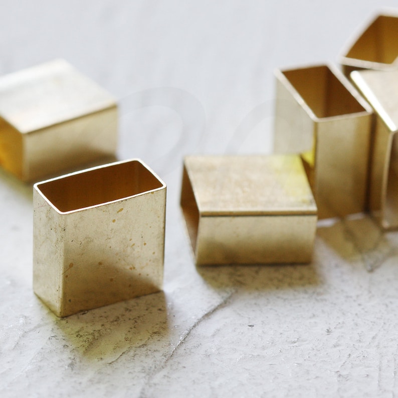 May include: A group of small, gold-colored metal squares with rounded edges. The squares are arranged in a loose pile on a white surface.