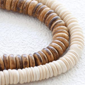 Może przedstawiać: Dwa sznury drewnianych koralików, jeden sznur jest brązowy, a drugi biały. Korale są okrągłe i mają lekko fakturowaną powierzchnię.