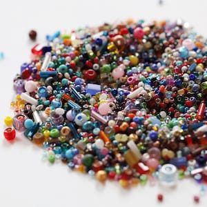 May include: A vibrant assortment of colorful beads in various shapes and sizes. The beads include round, cylindrical, and irregular forms in shades of red, blue, green, yellow, and pink. The beads are scattered on a white surface.