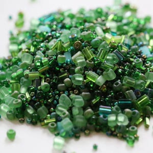 May include: A pile of assorted green beads in various shapes and sizes. The beads range in shades from light to dark green, including round, tube, and rectangular shapes. Some beads have a metallic sheen, creating a textured and visually appealing display, perfect for jewelry making.