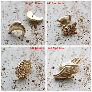 May include: Four pairs of gold-tone earring findings with different shapes, including a crescent moon, a croissant, a flower, and a pair of hands. The earring findings are displayed on a textured, light brown surface. #42 15x11mm, #43 16x10mm, #44 14mm, #46 25x13mm
