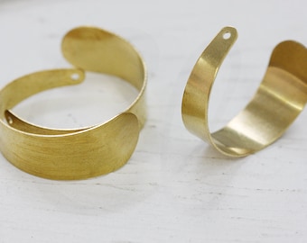 Pode incluir: Duas pulseiras de punho de metal dourado. As pulseiras têm um design curvo e aberto. Uma pulseira está fechada, enquanto a outra está aberta. Cada pulseira tem um pequeno orifício na extremidade da secção curva.