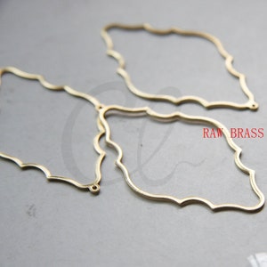 Könnte beinhalten: Zwei goldfarbene Metallscheiben für Ohrringe mit einem gewellten Rand. Die Scheiben haben die Form eines Rechtecks mit abgerundeten Ecken. RAW BRASS ist in Rot auf der Oberfläche aufgedruckt.