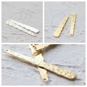 Puede incluir: Dos colgantes de barra de metal martillado, dos plateados y dos dorados, con dos agujeros en la parte superior. Los colgantes están sobre una superficie blanca.