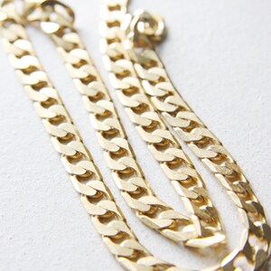 May include: A gold chain necklace with a thick, flat curb link design. The chain is folded in half and laid out on a white surface.