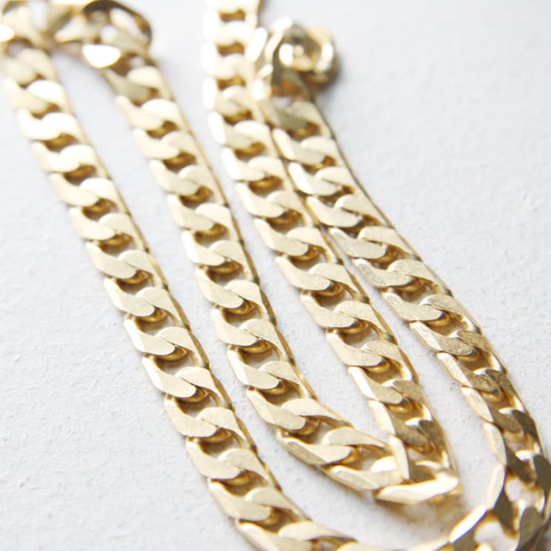May include: A gold chain necklace with a thick, flat curb link design. The chain is folded in half and laid out on a white surface.