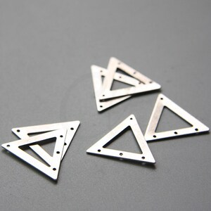 Puede incluir: Un conjunto de seis dijes de metal de forma triangular plateados con pequeños agujeros alrededor de los bordes. Los dijes están apilados uno encima del otro.